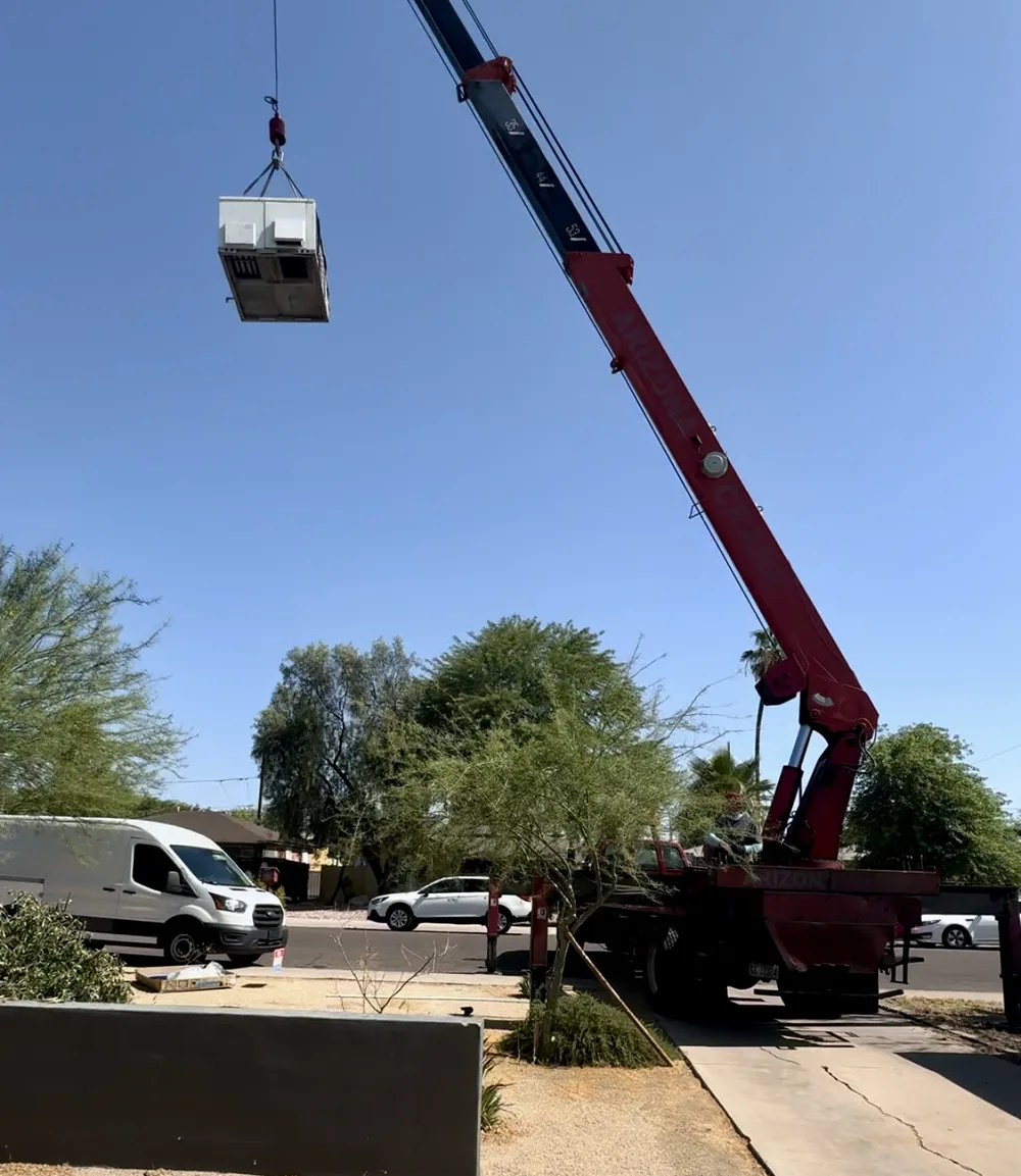 Boom crane parked on a residential street lifting a rooftop package AC unit over a desert-landscaped front yard
