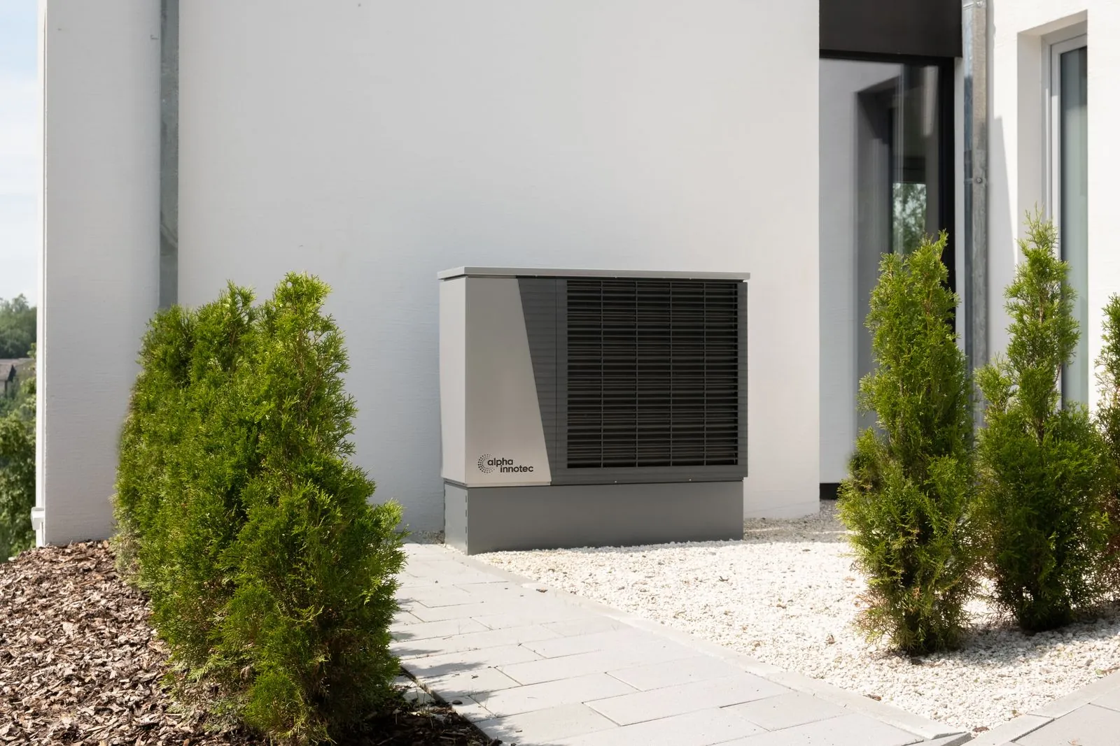 Outdoor heat pump unit on a paved path beside a white house, with shrubs on either side