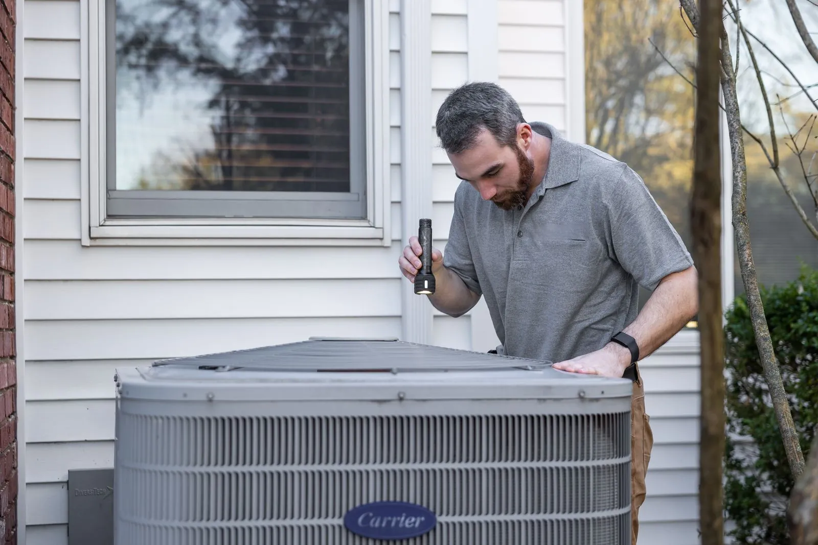 A man shining a flashlight over the top of an outdoor AC condenser beside a house